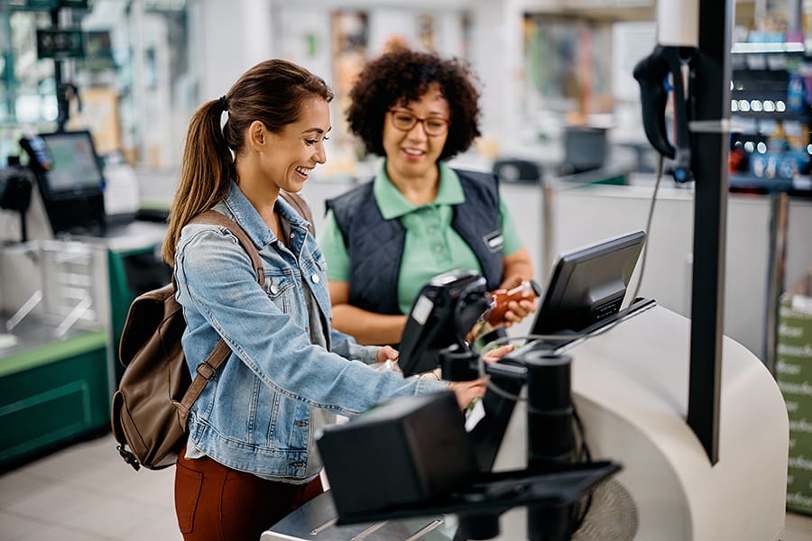 Young happy woman using self-service checkout with help of supermarket worker.