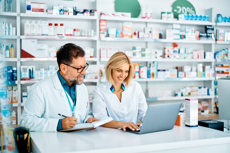 Happy pharmacist using laptop while her colleagues is taking notes in pharmacy.