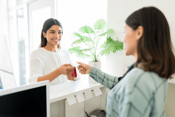 A smiling woman at a service counter hands a card to a customer standing across from her.